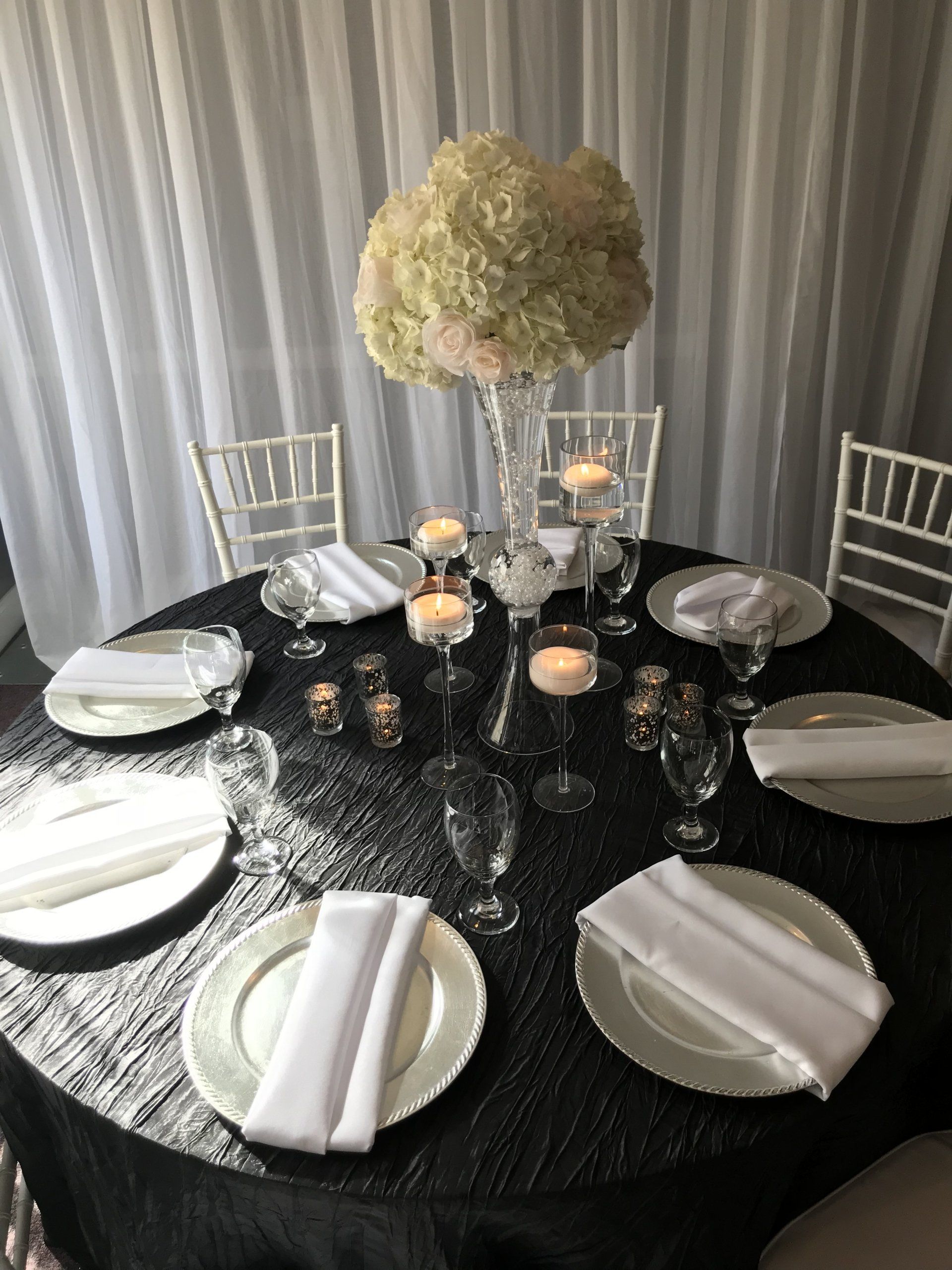 Table set up design with tall floral arrangement, various candles, silver chargers, and white napkins sitting on a crushed black tablecloth.