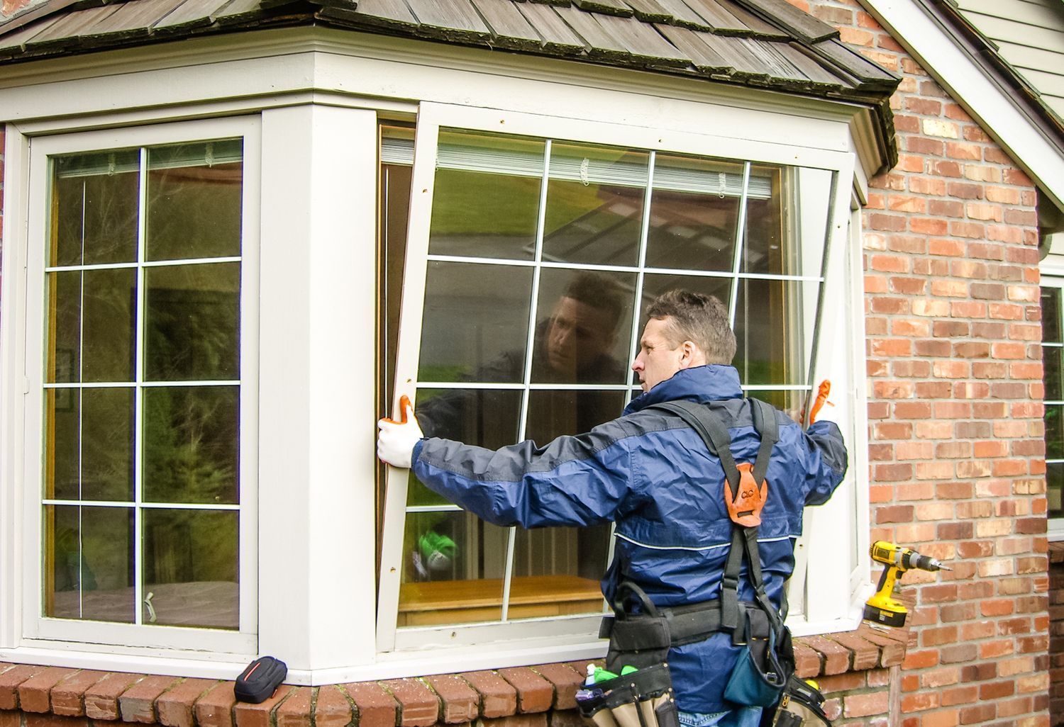 A worker in blue jumpsuit installing a window pane in a brick house. The window is white with a grid design.