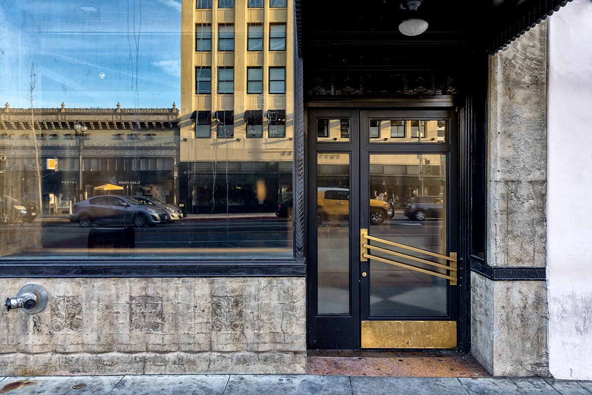 Exterior of a building with a black doorway, gold accents, and large glass windows. A car drives by, and other buildings are reflected.