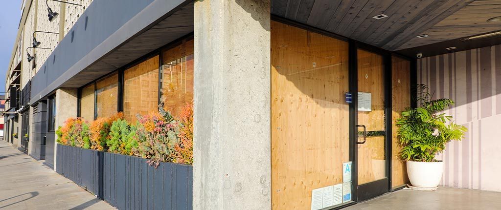Exterior of a commercial building with boarded-up windows and doors. A planter box holds colorful foliage along the sidewalk. A potted plant sits near the entrance.
