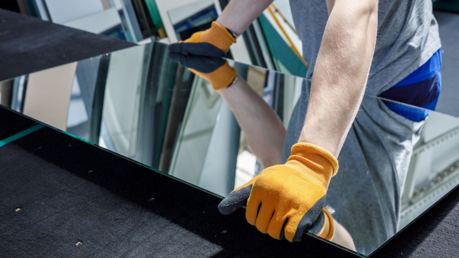 Person wearing orange gloves handling a large rectangular mirror in a workshop setting.