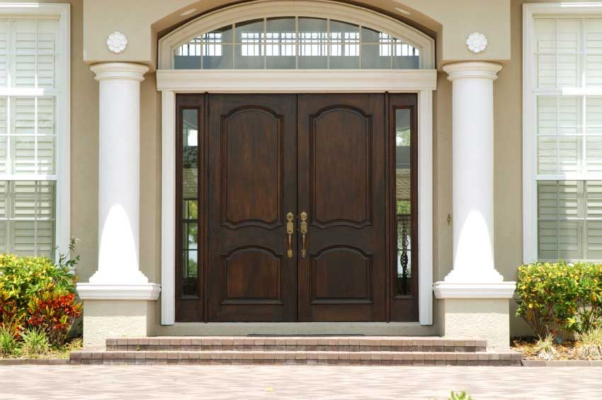 Double dark wood front doors with sidelights and arched window above, flanked by white pillars on a beige house.