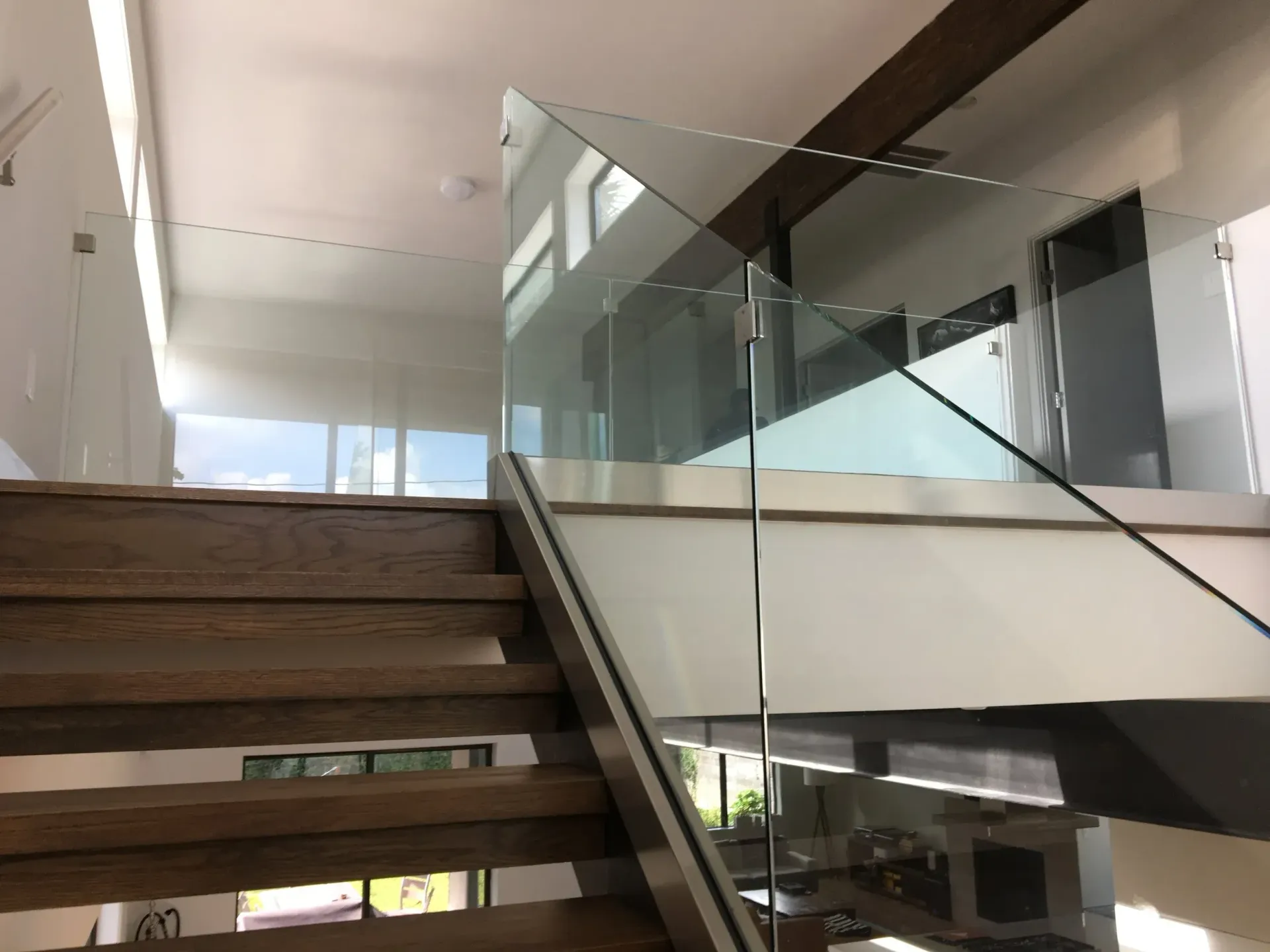 Wooden staircase leading up to a glass-railed landing in a modern home. Sunlight streams through windows.