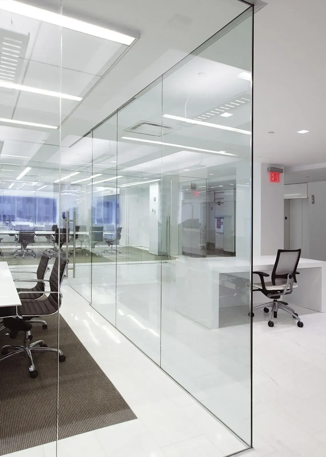 Modern office space with glass walls, white floors, and workstations. Black office chairs and an exit sign are visible.