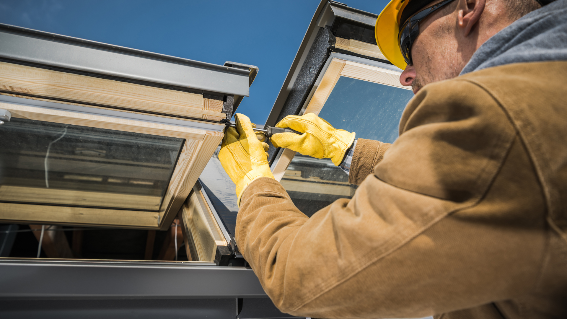 A construction worker in a hard hat and gloves repairs a skylight on a rooftop under a clear blue sky.