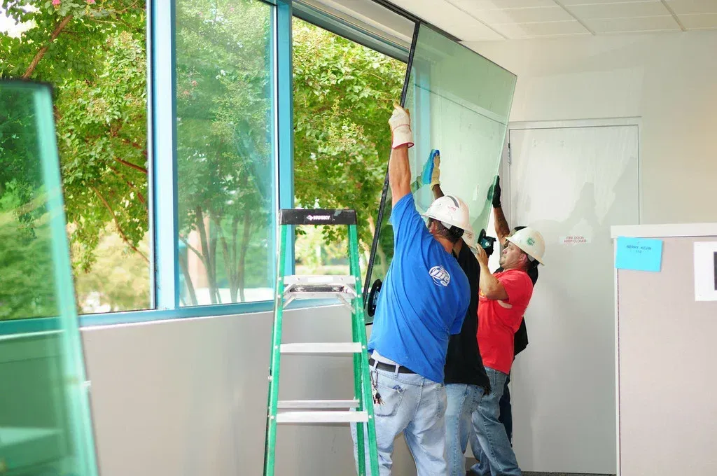 Two construction workers installing a large glass panel in an office, using a ladder.