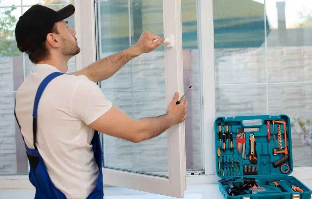 A window installer, in a white shirt and blue overalls, working on a white window with tools in a teal case nearby.
