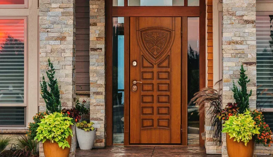 Wooden front door with decorative carvings, flanked by stone walls and potted plants.