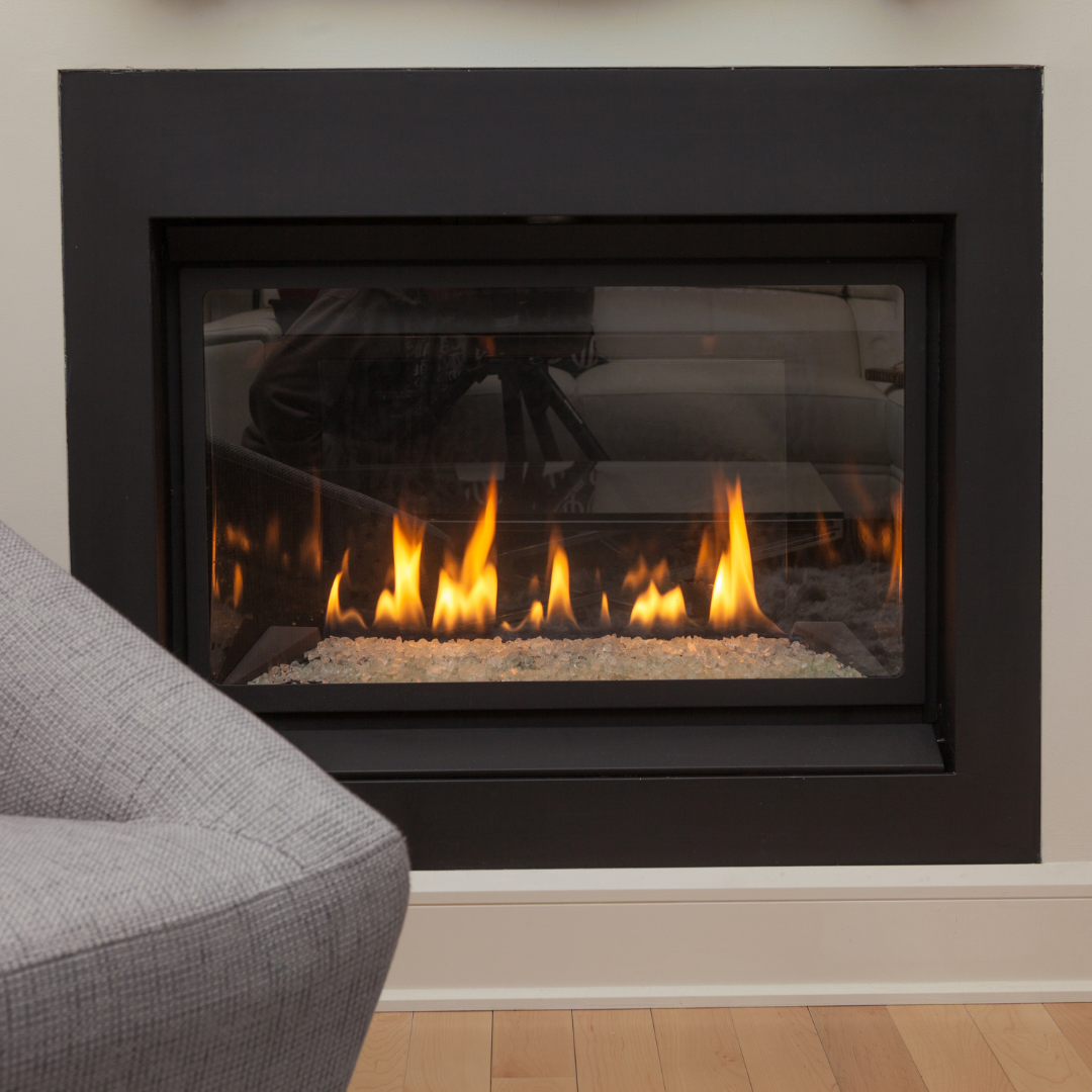 A modern, black-framed fireplace with a glass front, flames burning, and white pebbles at the base; a gray armchair is in the foreground.