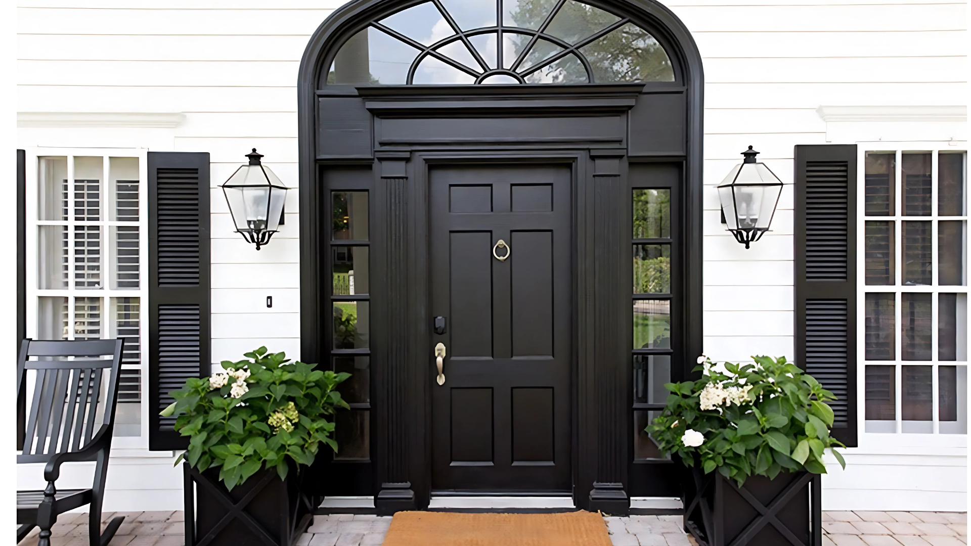 Black front door with arched transom window, flanked by sidelights, lanterns, and planters; set against a white facade.