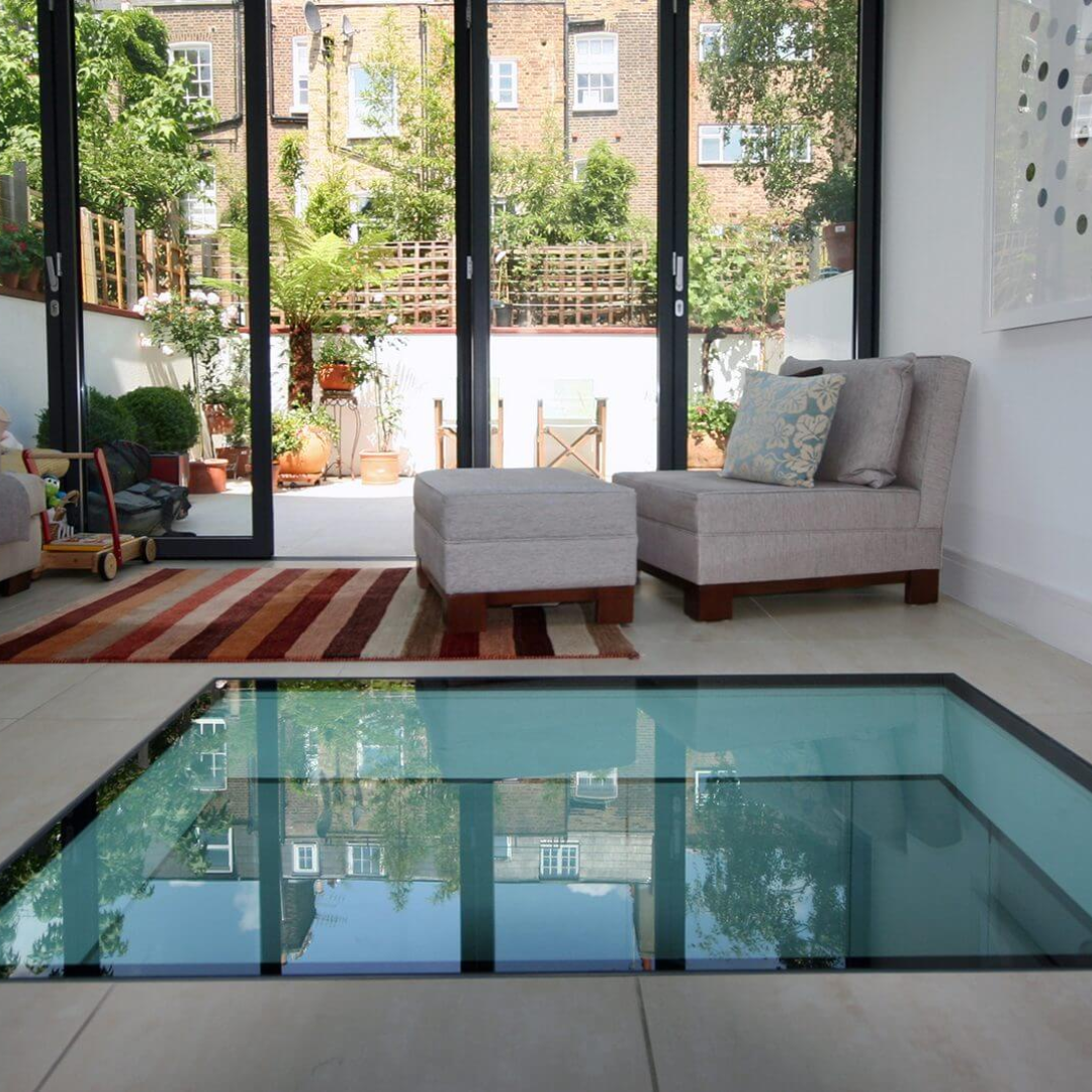 Living room with a glass floor, open to a patio.  Neutral furniture sits on a rug, while reflections of buildings are seen through the glass.