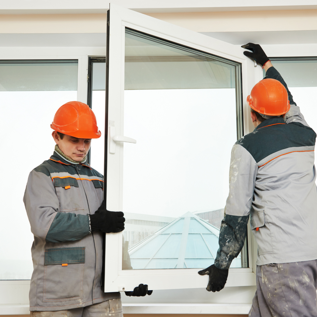 Two construction workers in orange hard hats installing a white window. They wear grey and teal work uniforms.