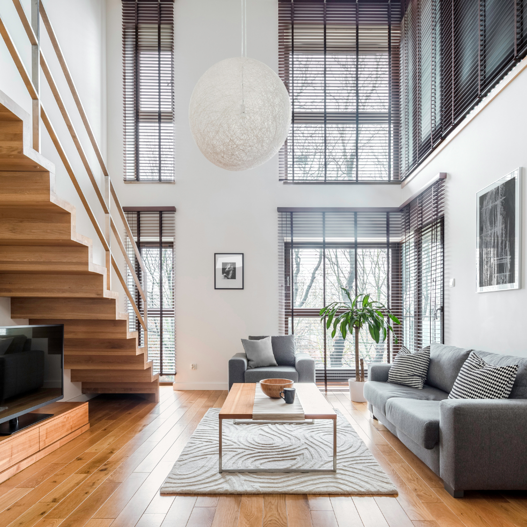 Spacious living room with wooden floors, stairs, and high windows with blinds. Gray sofas, coffee table, and a large hanging light fixture.