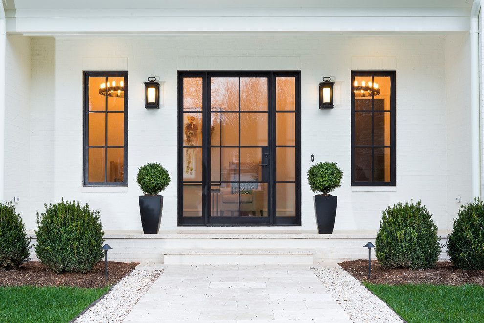 White house entrance with black-framed door and windows, featuring symmetrical design elements, two planters, and exterior lights.