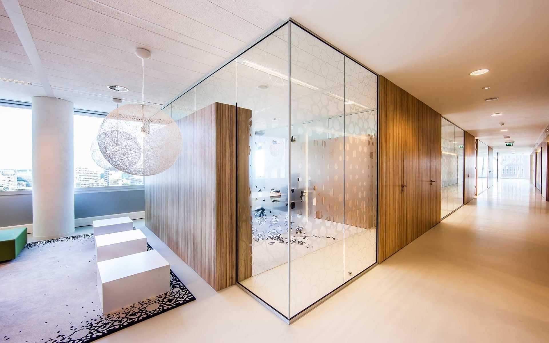 Modern office interior with glass-walled meeting room, wooden paneling, and bright hallway. A decorative hanging light and outdoor view are visible.