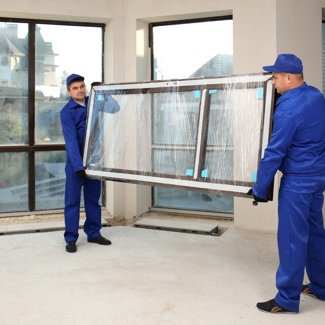 Two workers in blue overalls carry a wrapped window indoors. They are lifting it across a light, unfinished room.
