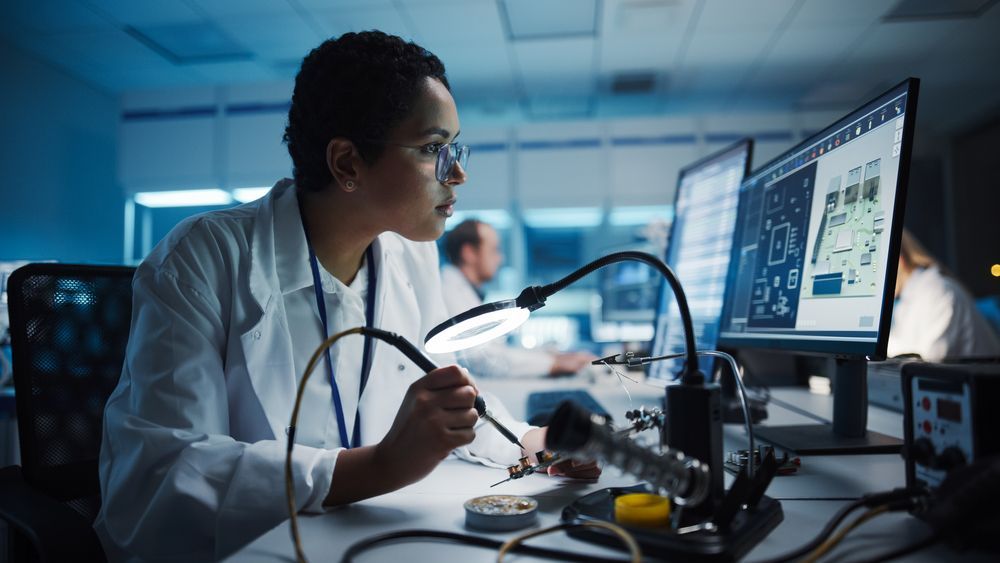 Woman in lab coat soldering circuit board at a workstation, looking at a computer screen.