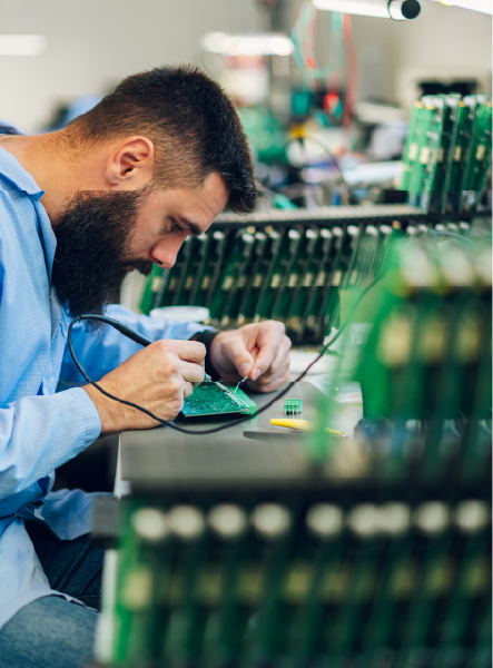 Man soldering circuit board in a lab setting, surrounded by rows of green boards.