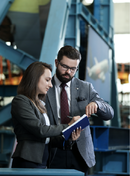 Woman and man in suits reviewing documents in a factory setting; man pointing, woman looking.
