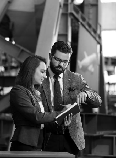 A man and woman in business attire review a notebook in a large industrial space.