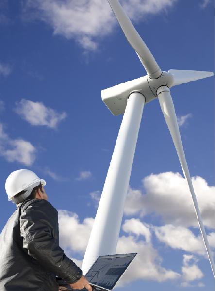 Engineer in hard hat examines wind turbine, laptop in hand, against a blue sky with clouds.