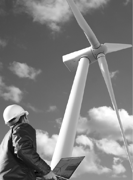 Engineer in hard hat working on laptop, inspecting wind turbine against cloudy sky.