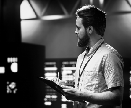 Man with a beard writing on a clipboard in a control room, surrounded by monitors.