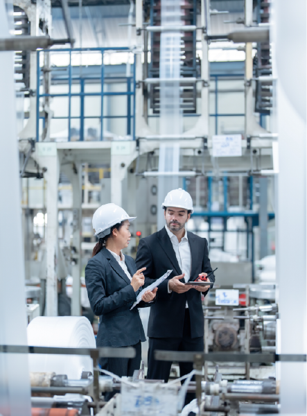 Two people in hard hats and suits inspect machinery in a factory.