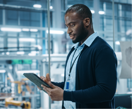 Black man in a factory, looking at a tablet. Wearing blue sweater, shirt, and lanyard.