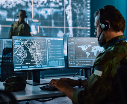 Soldier in command center, using computers with maps and data, wearing headset, other soldier in background.