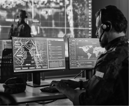 Two military personnel in a control room, working at computers with maps and data, wearing headsets.