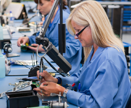 Two women in blue lab coats soldering circuit boards at a workbench in a manufacturing setting.