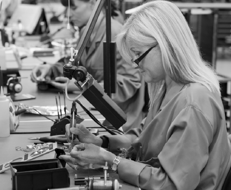 Woman soldering components at a workstation; another worker is in the background.