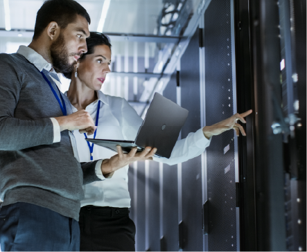 Two IT professionals inspecting server racks in a data center; one pointing, one holding a laptop.