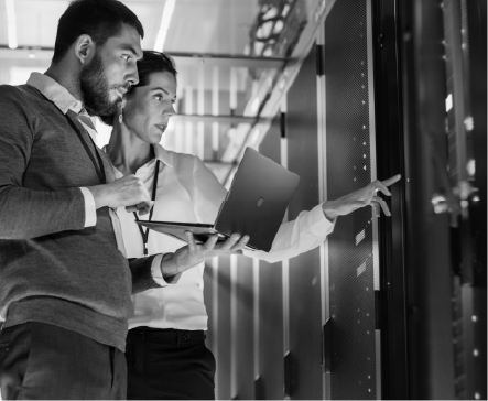 Two IT professionals in a server room, examining equipment. Woman points, man holds a laptop.