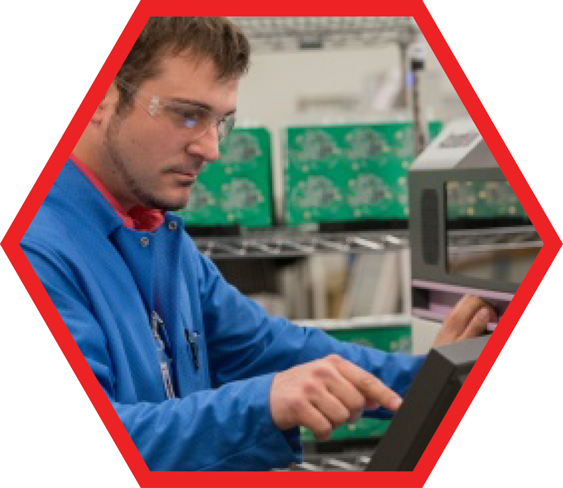 Man in blue lab coat and safety glasses, operating a machine with circuit boards in the background.