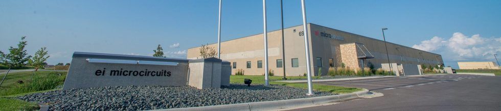 Exterior view of a tan industrial building and a sign on a small cement structure that reads