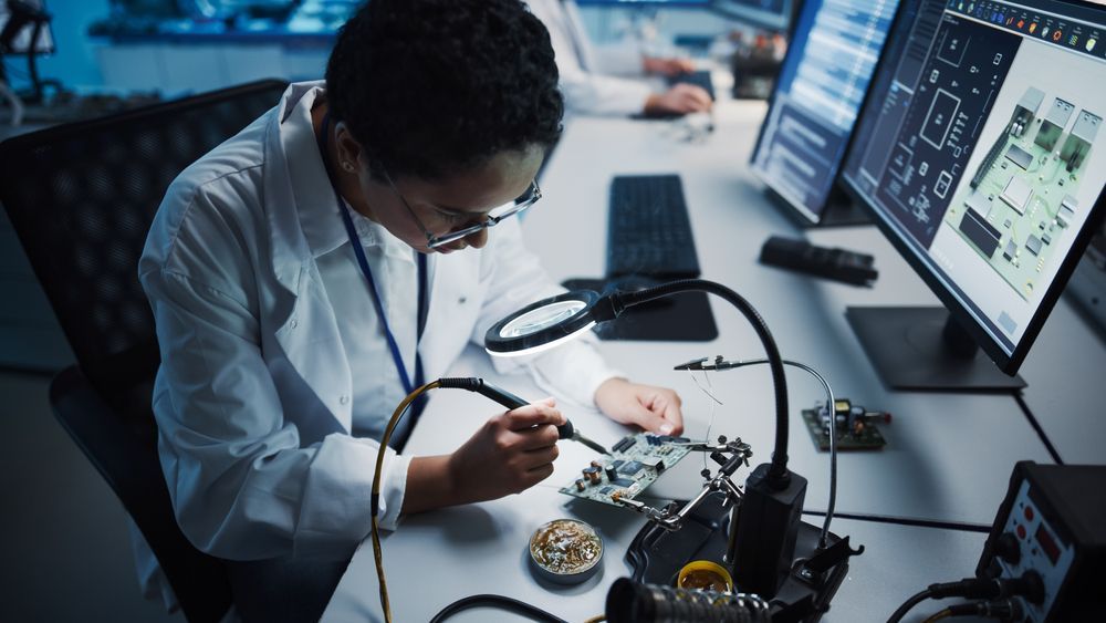 Woman soldering circuit board in lab with magnifying glass and computer screens.