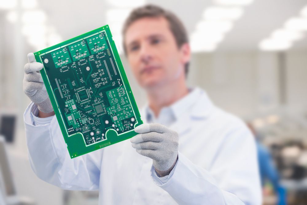 Man in lab coat holding a green circuit board, inspecting it.