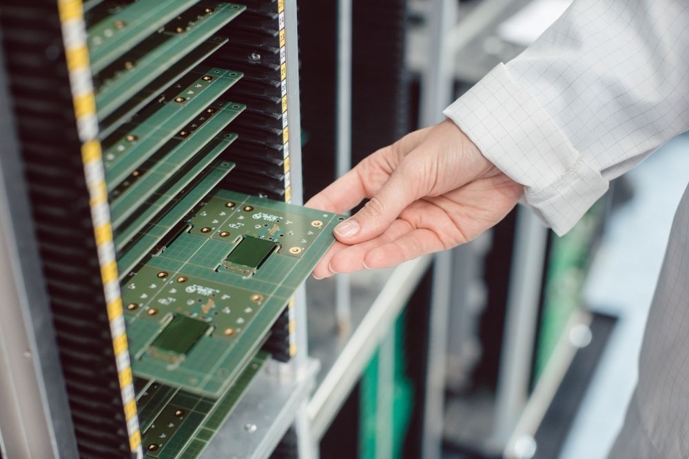 A person in a lab coat removes a green circuit board from a rack in a laboratory setting.