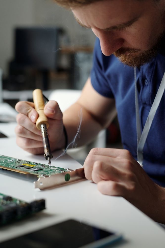 Man soldering electronic circuit board at a white table. He's holding a soldering iron and wire, focused intently.