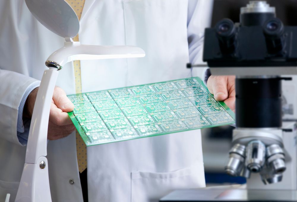 Person in a lab coat examines a green printed circuit board under a magnifying lamp.