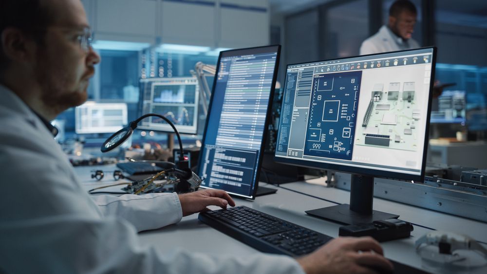 Man in lab coat working at computer with blueprints and code; laboratory setting.