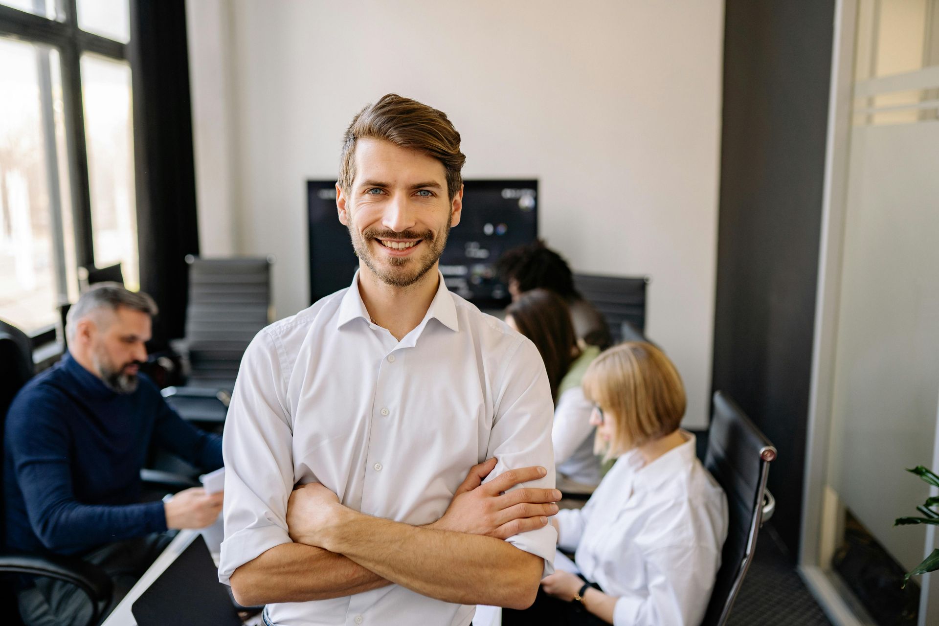 A man is standing with his arms crossed in front of a group of people in an office.