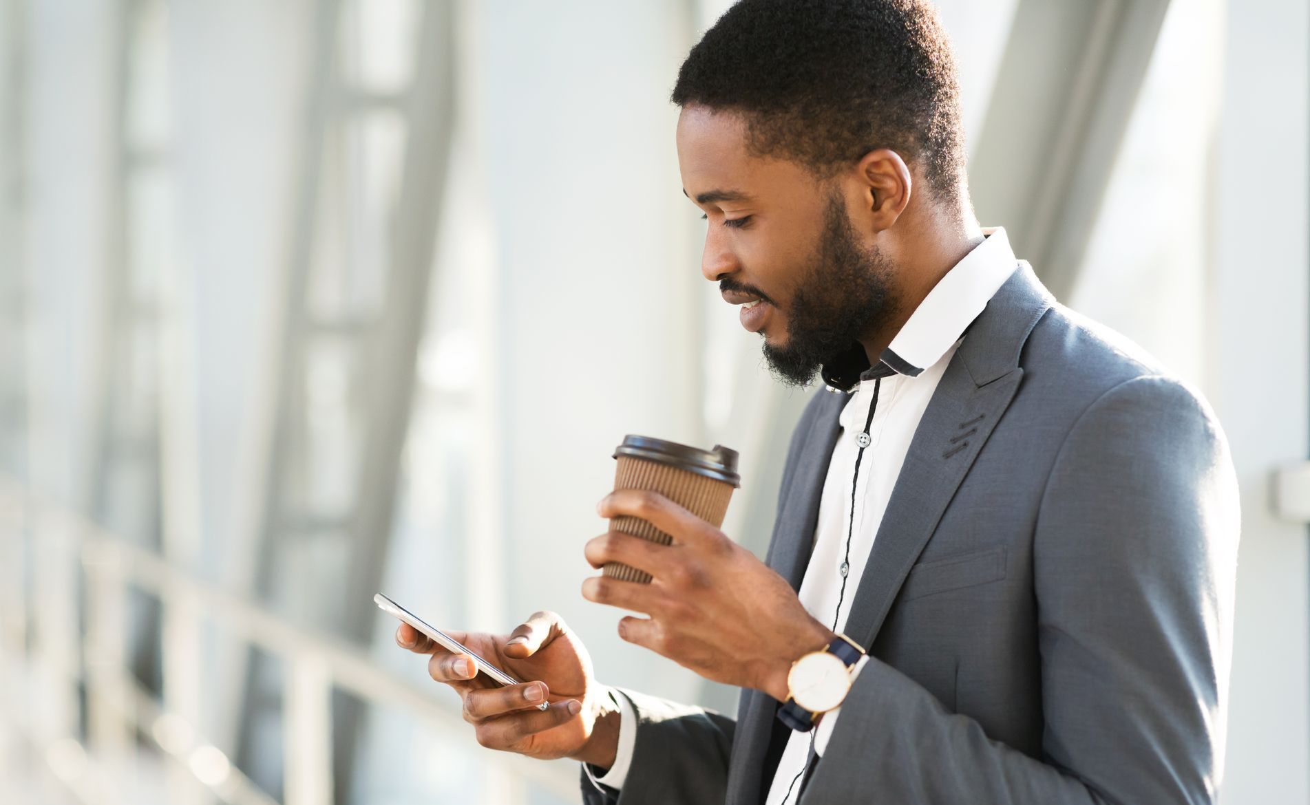 Man in a suit holding a cup of coffee and looking at his cell phone.