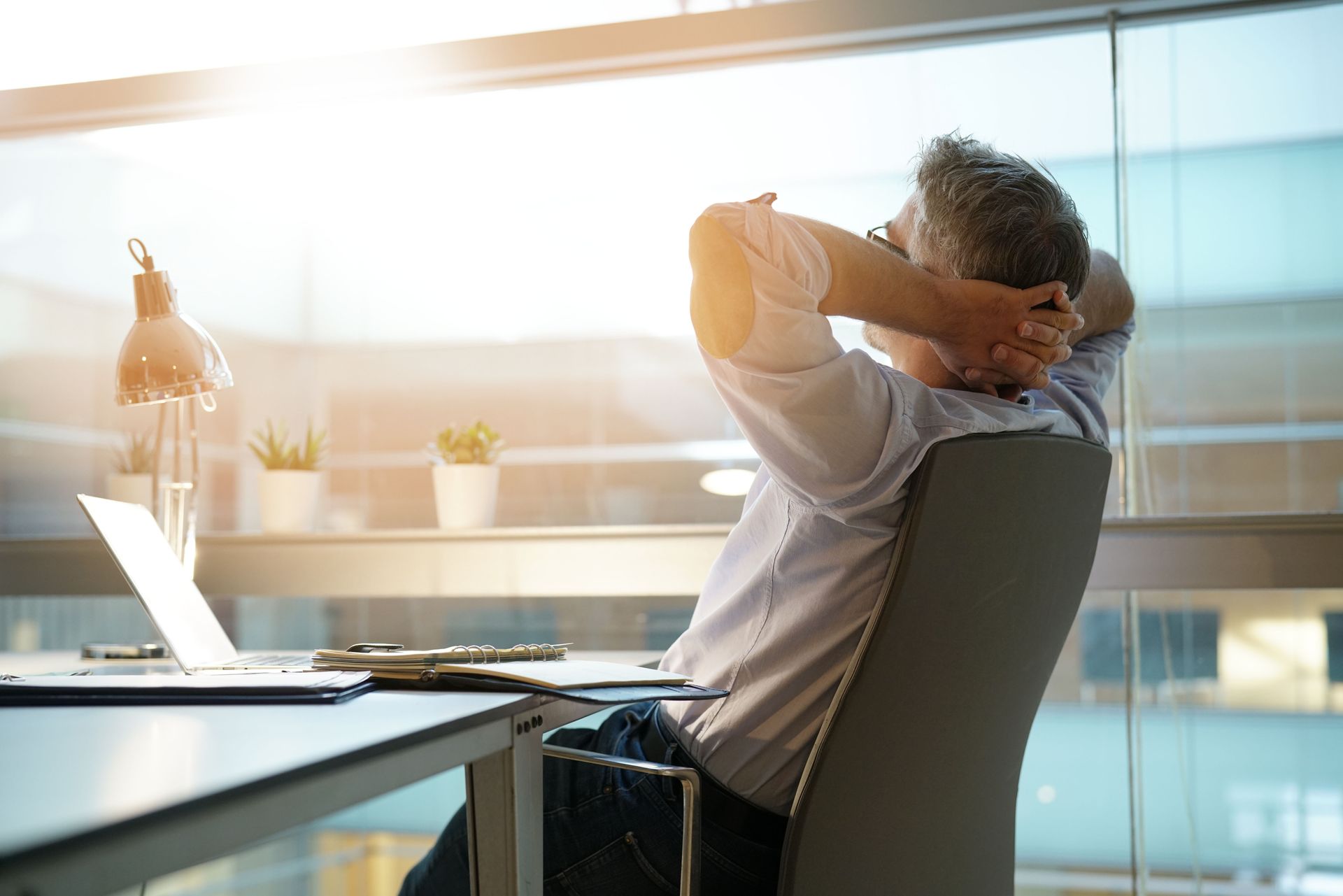 Man relaxing at a desk in an office, hands behind head, overlooking a sunny cityscape.