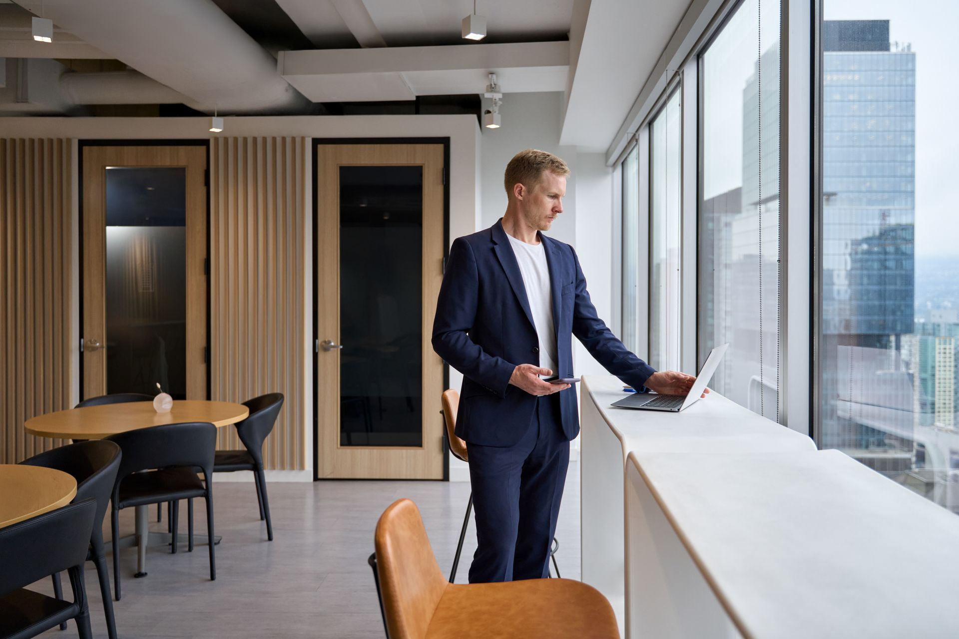 Man in a suit using a laptop by a window overlooking a cityscape.