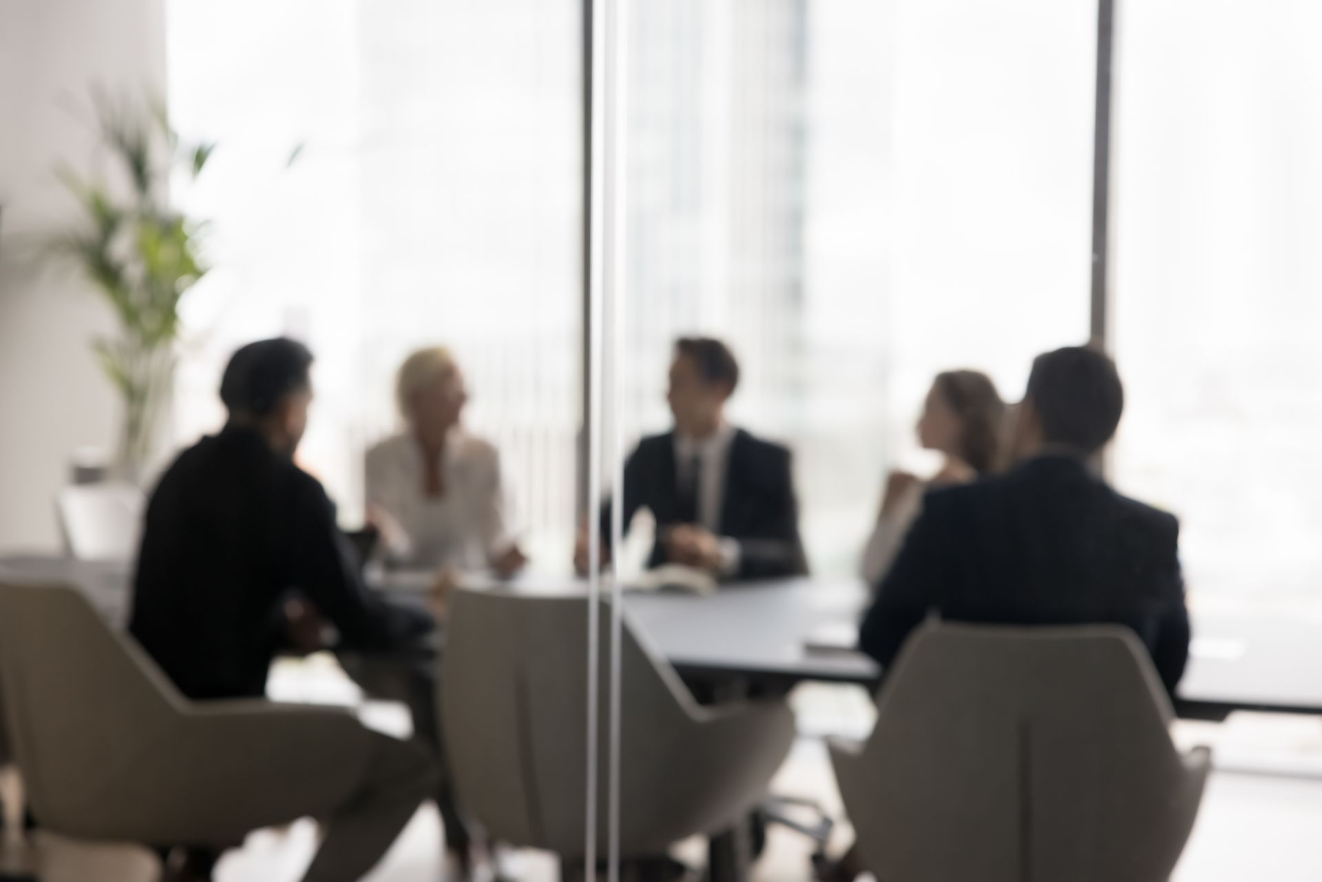 Blurred view of a business meeting in a modern office with glass walls; people seated around a table.