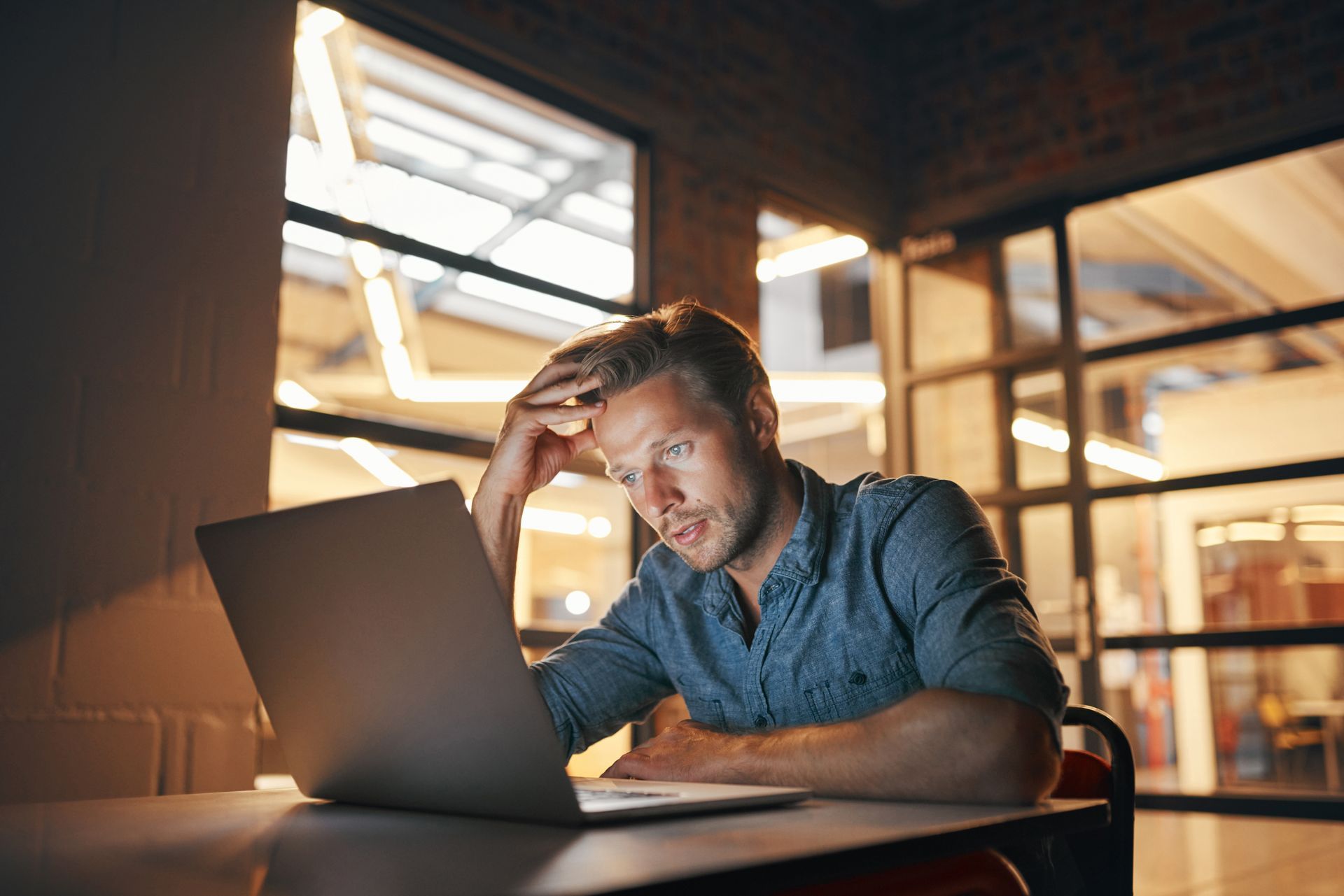Man with hand on forehead, looks stressed at laptop in low light.