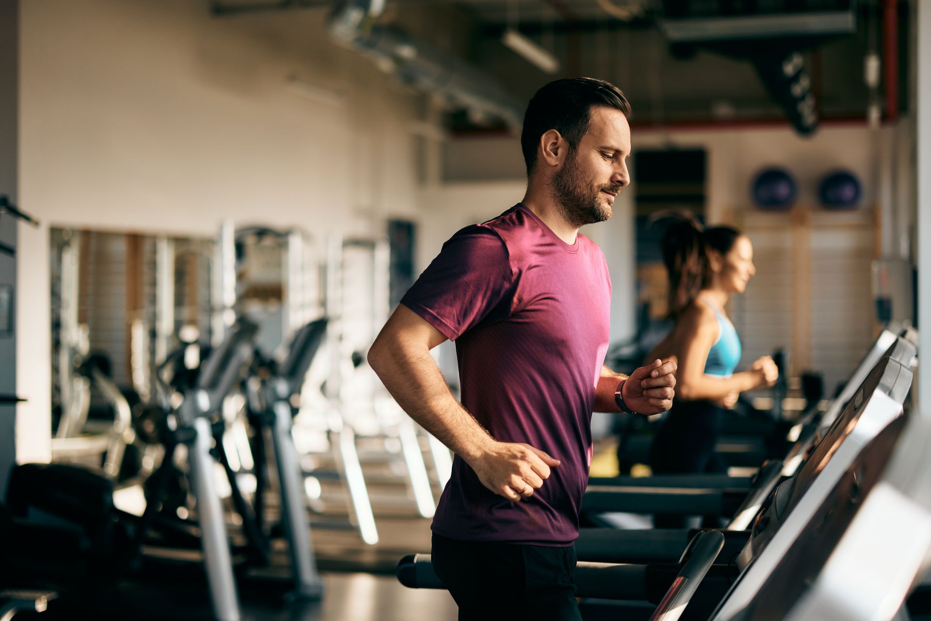 Man in a purple shirt running on a treadmill at a gym, another person behind him.